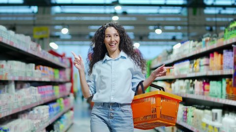 cheerful young woman customer in the store dances funny between the rows in a supermarket with a basket in his hands. walking the grocery store and moving like dancing while doing shopping having fun, videoclip de stoc