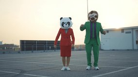Cinematic video of a couple wearing elegant suits and funny animals masks having fun and celebrating outdoor in a a parking lot rooftop	
