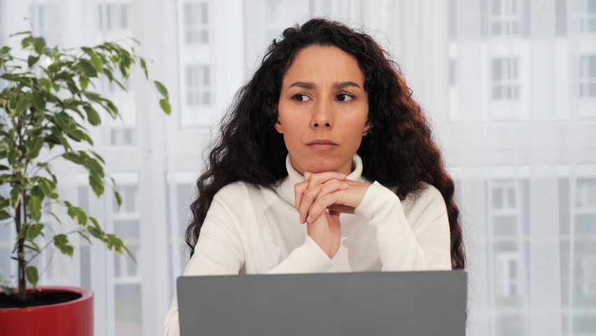Thoughtful serious young hispanic curly hair woman student writer sit at home office with laptop thinking of inspiration search problem solution ideas lost in thoughts concept dreaming looking away. Royalty-Free Stock Footage #1096521369