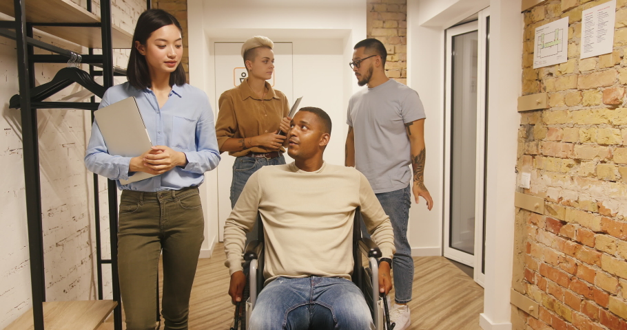 Disabled black man drives wheelchair talking to colleagues walking in office corridor. International co-workers discuss joint project slow motion Royalty-Free Stock Footage #1096528609