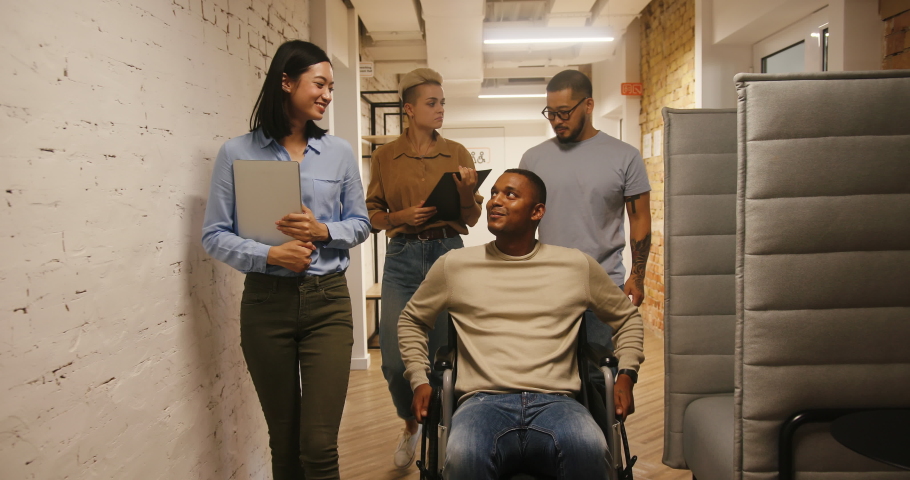 Disabled black man drives wheelchair talking to colleagues walking in office corridor. International co-workers discuss joint project slow motion | Shutterstock HD Video #1096528609