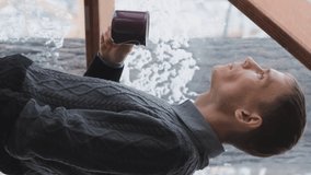 Vertical video. Young man drinking hot chocolate or cocoa and looks out of window of dome camping, enjoying scenic view of snow-covered winter nature, slow motion