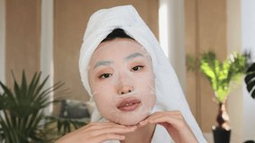 Portrait of beautiful young asian woman with cotton mask sheet on face and towel on head over exotic background. Pretty lady doing skin care procedures indoors at home. - Powered by Shutterstock - Get 15% off with code: PIKWIZARD15
