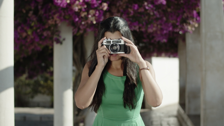 Front view of young Hispanic woman taking photo with camera, helping friend with choosing best posture while enjoying warm summer day, making shots. Camera POV. Photography, travel concept.
