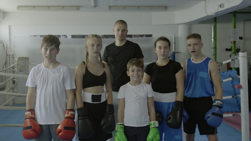 Boxing class kids and teacher showing winner gesture portrait, raising hands in gloves, front view medium shot. Children and boxer looking at camera, standing in gym