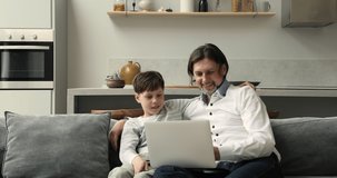 Happy foster dad adopted son teenager cuddle on couch focused on laptop screen watch video read sport news play game. Caring father software engineer teach tween boy to make simple program code on pc
