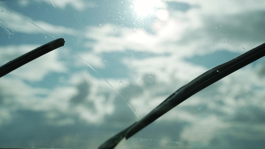 Wiper blades remove raindrops from the windshield of the car while driving, close-up, handheld shot. End of rain, drops of water fall on the glass, sun shines through the clouds, view of the sky. | Shutterstock HD Video #1096540935
