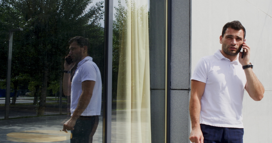 Businessman talking on smart phone. Male professional is reflecting on glass window while standing against building. He is socializing in city.