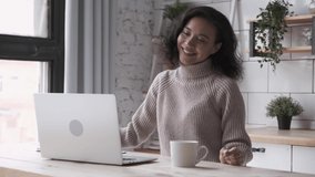 Black lady has online video call with friends telling about autumn weather. Young joyful woman sits at kitchen table gesturing with hands at laptop screen