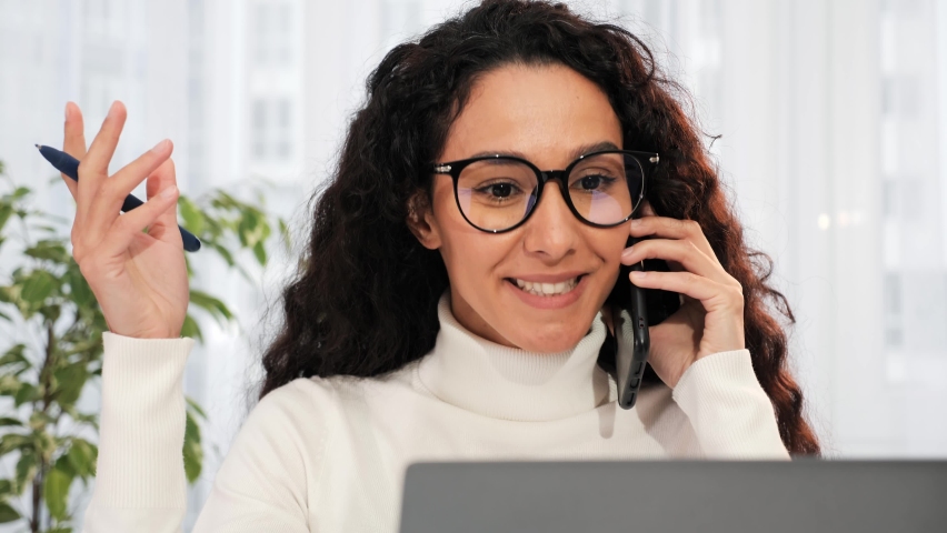 Smiling young business woman professional in glasses talking on phone using laptop sit at home office desk, happy female customer make mobile call confirming online website shopping order delivery. Royalty-Free Stock Footage #1096553127