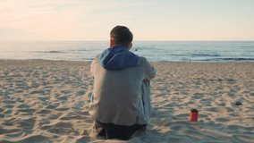 Back view of young Caucasian man sitting on sandy beach, relaxing, enjoying sea view and drinking coffee. Summer vacation concept