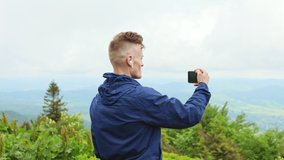 Back view of young, sporty male with earphones hiking, taking photo of hills. Male wearing sportswear, standing on hill, admiring beauty of nature. Concept of traveling and hiking.
