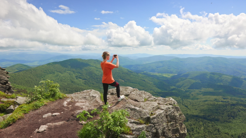 Side view of sporty woman wearing T-shirt, holding phone, taking photo. Slim female tourist climbing hill, standing on rock, looking forward. Concept of traveling and hiking. Royalty-Free Stock Footage #1096556025