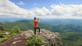 Side view of sporty woman wearing T-shirt, holding phone, taking photo. Slim female tourist climbing hill, standing on rock, looking forward. Concept of traveling and hiking.