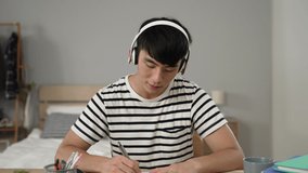 concentrated asian male college student looking at webcam with headphones is nodding head and taking notes while learning from home taking online lesson