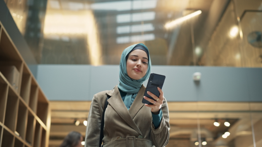 Muslim Woman wearing Hijab Checking Email App on her Smartphone After a Succesful Meeting. Portrait of a Female Content Manager Walking Through a Busy Workplace Hall. Low Angle