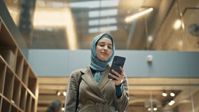 Muslim Woman wearing Hijab Checking Email App on her Smartphone After a Succesful Meeting. Portrait of a Female Content Manager Walking Through a Busy Workplace Hall. Low Angle - Powered by Shutterstock - Get 15% off with code: PIKWIZARD15