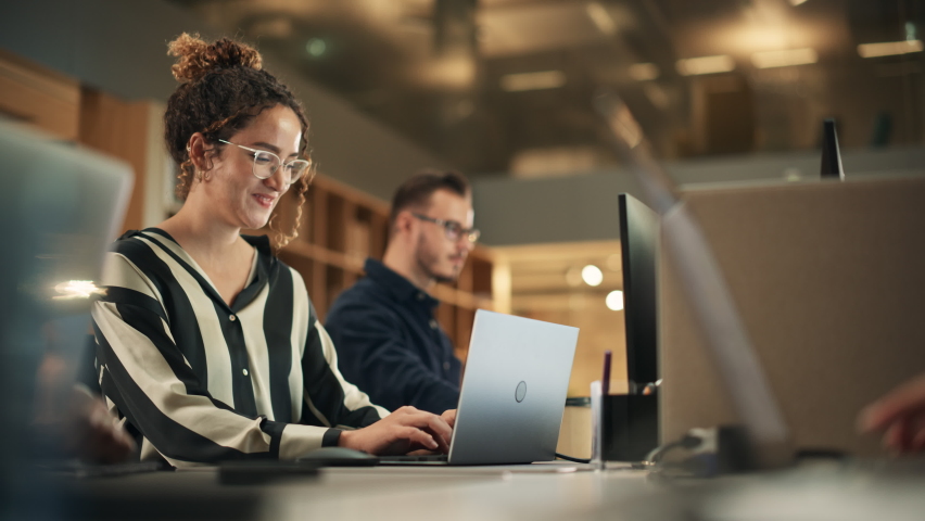 Portrait of Enthusiastic Hispanic Young Woman Working on Computer in a Modern Bright Office. Confident Human Resources Agent Smiling Happily While Collaborating Online with Colleagues. Dolly Shot