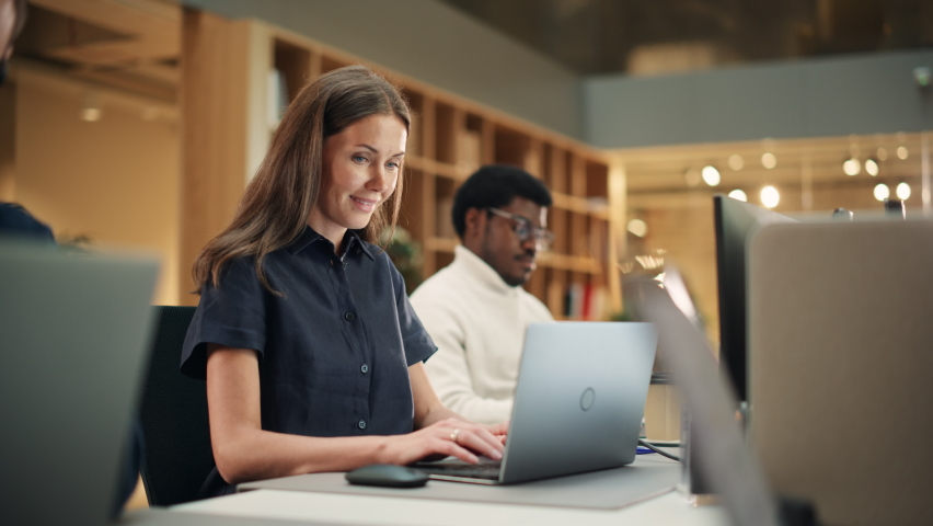 Multiethnic Group of Creative Colleagues Working on Laptops During Day in a Modern Bright Office. A White Woman Smiles Happily After Successfully Finishing a Work Project. Slow Motion