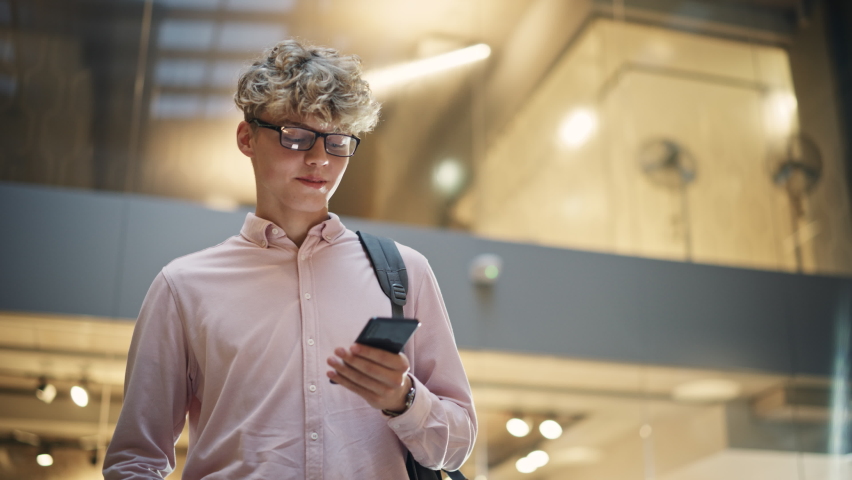 Portrait of University Student Checking his Text on his Smartphone and Smiling While he Walks Toward Classroom. Young White Man Male Crossing the Hall of a Big Building. Low Angle, Slow Motion Shot Royalty-Free Stock Footage #1096559171
