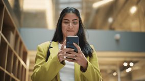 Portrait of a Happy Hispanic Woman Texting on her Smartphone and Walking in Corporate Office. Female Marketing Manager Checking Social Media Trends and Staying Updated. Low Angle Slow Motion - Powered by Shutterstock - Get 15% off with code: PIKWIZARD15