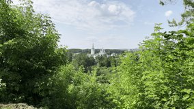 Green summer forest with city buildings behind. Clip. Blue cloudy sky and a small town behind trees.