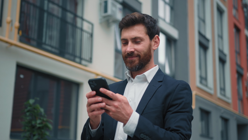 Attractive caucasian businessman stand in city texting messages on mobile phone positive smiling 30s freelancer owner read internet digital news good notification browsing media content online working Royalty-Free Stock Footage #1096561093