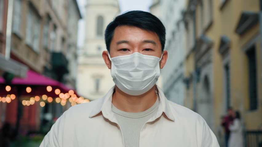 Headshot adult asian man in protective medical mask stands in the city outdoors taking off respirator take deep breath fresh air healthy happy male tourist enjoying end of quarantine looking at camera Royalty-Free Stock Footage #1096561151