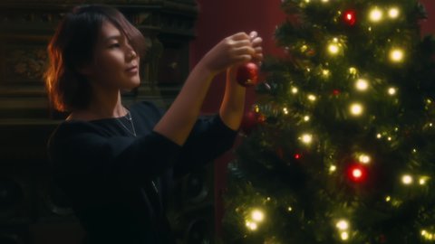 Young asian woman hangs red shiny ball on Christmas tree at home. Attractive female brunette puts toys and garlands on the Christmas tree on the of New Year eve celebration - Βίντεο στοκ