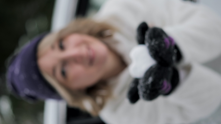Happy woman in stylish winter clothes holding heart shape snow ball. Looking out of car window. Valentine