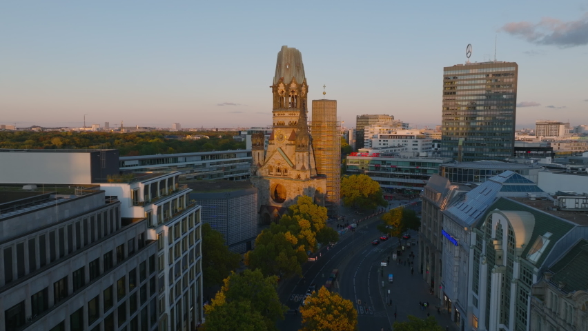Forwards fly above street in modern urban borough. Heading towards remains of Kaiser Wilhelm Memorial Church lit by setting sun. Charlottenburg neighbourhood, Berlin, Germany