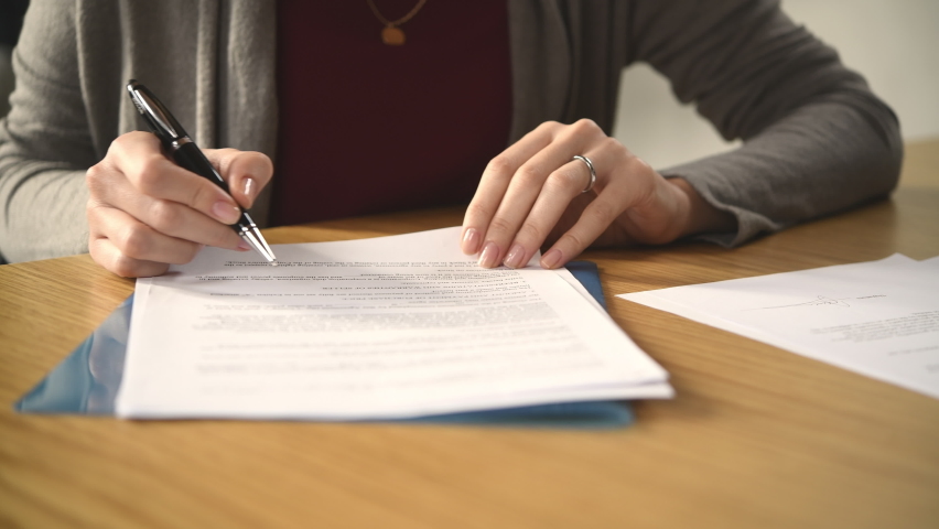 Close up of hands of young woman signing legal documents on desk. Close up of woman hand holding pen and signing legal paper seated at desk. Detail of lawyer filling official document, deal done. Royalty-Free Stock Footage #1096566045