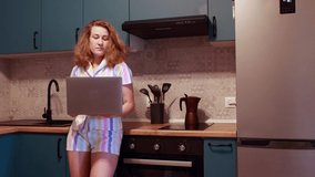 A young woman is standing in the kitchen in pajamas. shifts a sad and thoughtful look away from the laptop.