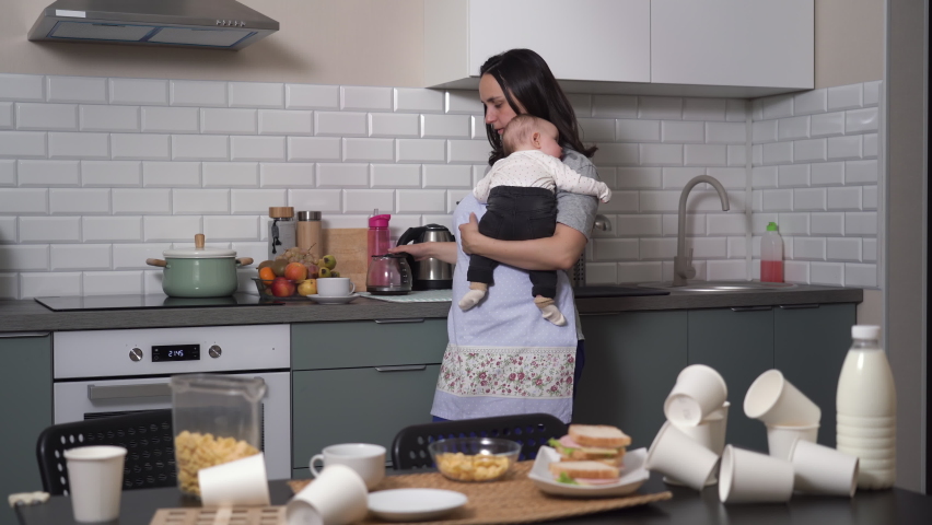 Exhausted young woman drinking coffee while holding baby in messy kitchen, tired mother lulling the child | Shutterstock HD Video #1096571415