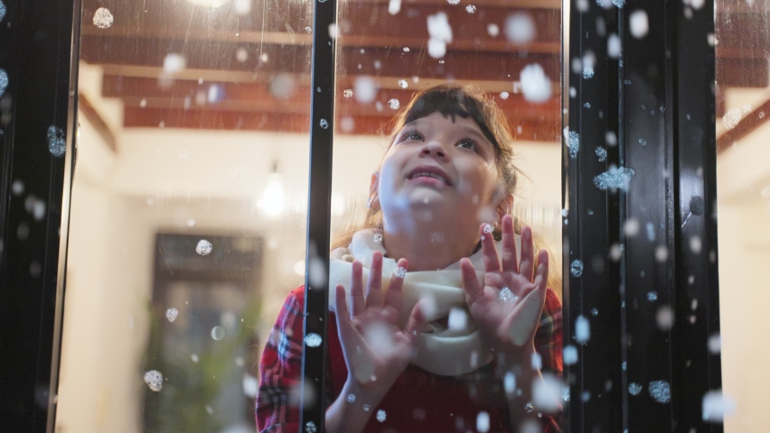 Adorable child look through the window and admiring first snow flakes. Young kid girl sitting in living room at home, watching snow falling outside enjoy celebrate Christmas, Thanksgiving with family.
