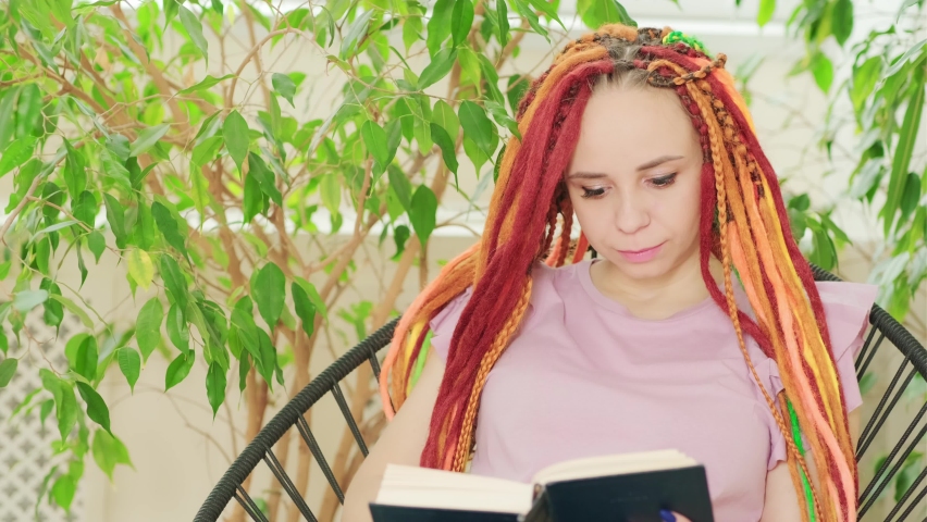 Young woman with dreadlocks reading book, sitting in chair surrounded by houseplants.