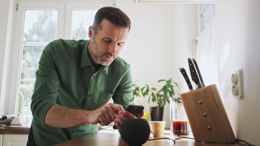 Adult man setting up smart speaker using smartphone in kitchen