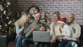 Family celebrates Christmas and New Year inside house with Christmas tree and gifts. Men and women make video call to relatives and friends on laptop and happily communicate congratulations holiday