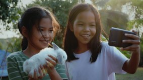 Two asian girls taking a picture and playing with baby bunny. Sister siblings holding a little rabbit Holland lop on hand and using smartphone selfie together with fun in the yard at the sunset time.