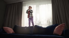 Slow motion video of Caucasian boy jumping on couch in living room. The concept of alone at home is a happy carefree childhood, the child fools around and rejoices. High quality 4k footage