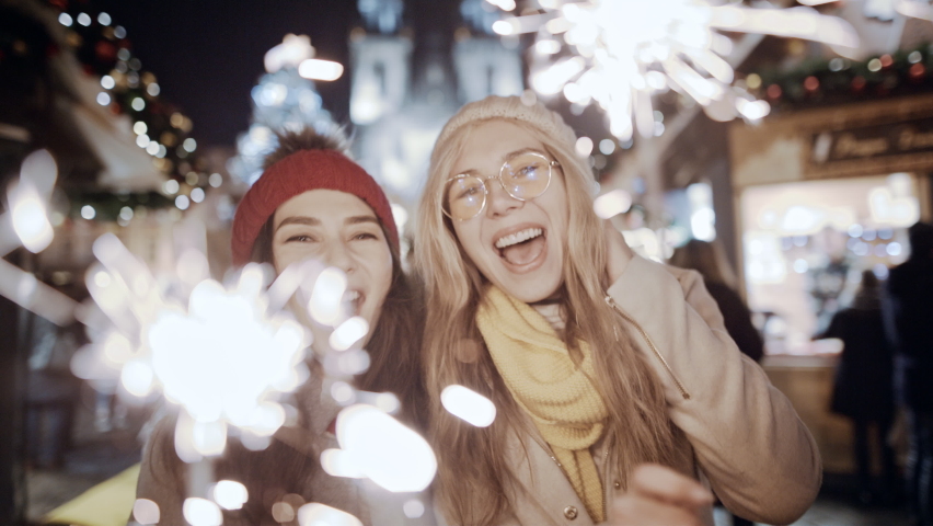 Celebrating New Year holidays vacation. Happy female friends with sparklers smile at decorated town square. Xmas Christmas winter atmosphere. Excited girls people holding lights in waving hands.