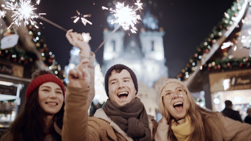 Celebrating New Year holidays Christmas vacation. Happy dancing three friends with sparklers smile at decorated town square. Xmas Christmas winter night atmosphere. Excited people take selfie on phone | Shutterstock HD Video #1096609229