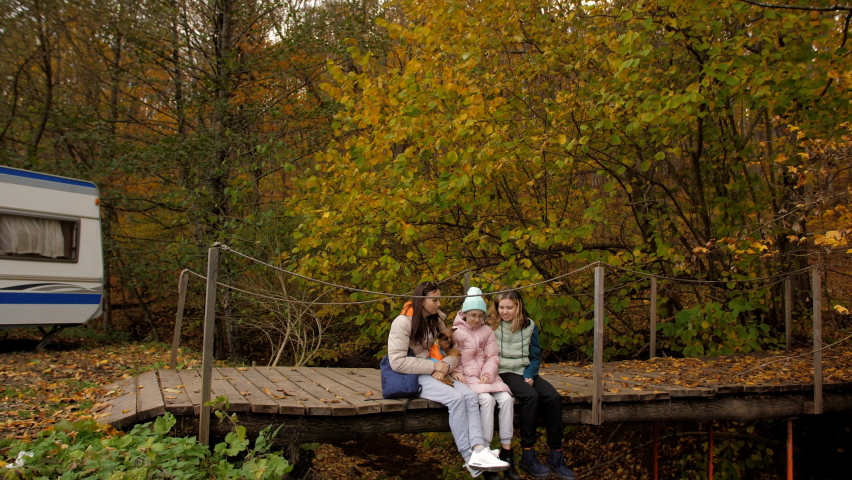 A woman and her two daughters are sitting on a wooden bridge over the river with their legs dangling, not far from a forest path. Autumn vacation in the forest.