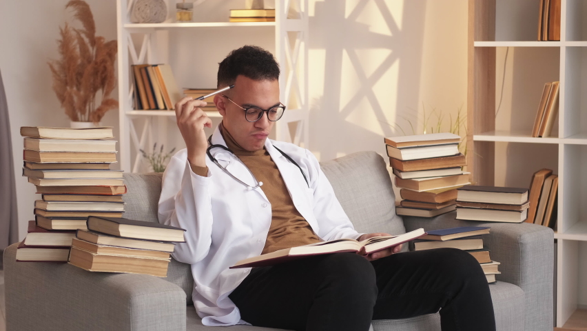 Learning medicine. Doctor training. Professional skill development. Pensive smart male intern studying thinking reading on couch with book pile at modern home interior. Royalty-Free Stock Footage #1096616475