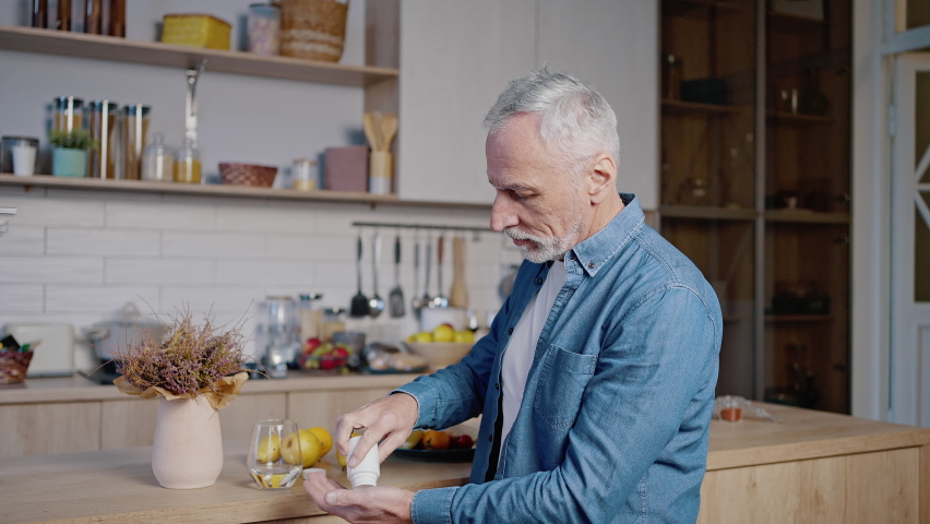Old man wearing blue shirt takes medicine. Grey-haired gentleman takes out pill from bottle on palm and drinks with water standing in kitchen Royalty-Free Stock Footage #1096620013