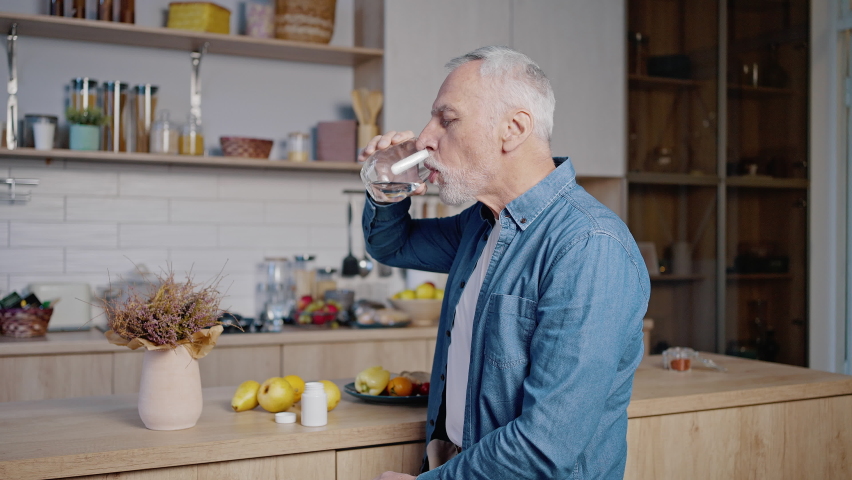Old man wearing blue shirt takes medicine. Grey-haired gentleman takes out pill from bottle on palm and drinks with water standing in kitchen Royalty-Free Stock Footage #1096620013