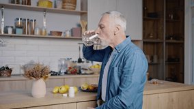 Old man wearing blue shirt takes medicine. Grey-haired gentleman takes out pill from bottle on palm and drinks with water standing in kitchen - Powered by Shutterstock - Get 15% off with code: PIKWIZARD15
