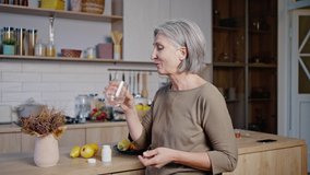 Elderly woman stands in light kitchen and takes medicine. Gray-haired female person takes out pill from bottle and drinks with water smiling - Powered by Shutterstock - Get 15% off with code: PIKWIZARD15