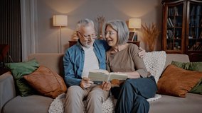 Elderly married couple sits on sofa in cozy living room. Couple hugs lovingly and reads book together. Husband and wife laugh happily discussing story - Powered by Shutterstock - Get 15% off with code: PIKWIZARD15
