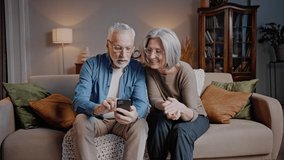 Couple of elderly people looks at mobile phone sitting on sofa in light living room. Happy man and woman laugh having pleasant conversation at home - Powered by Shutterstock - Get 15% off with code: PIKWIZARD15